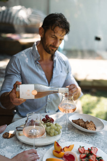 Man pouring wine at a outdoor dining table with food and fruit.